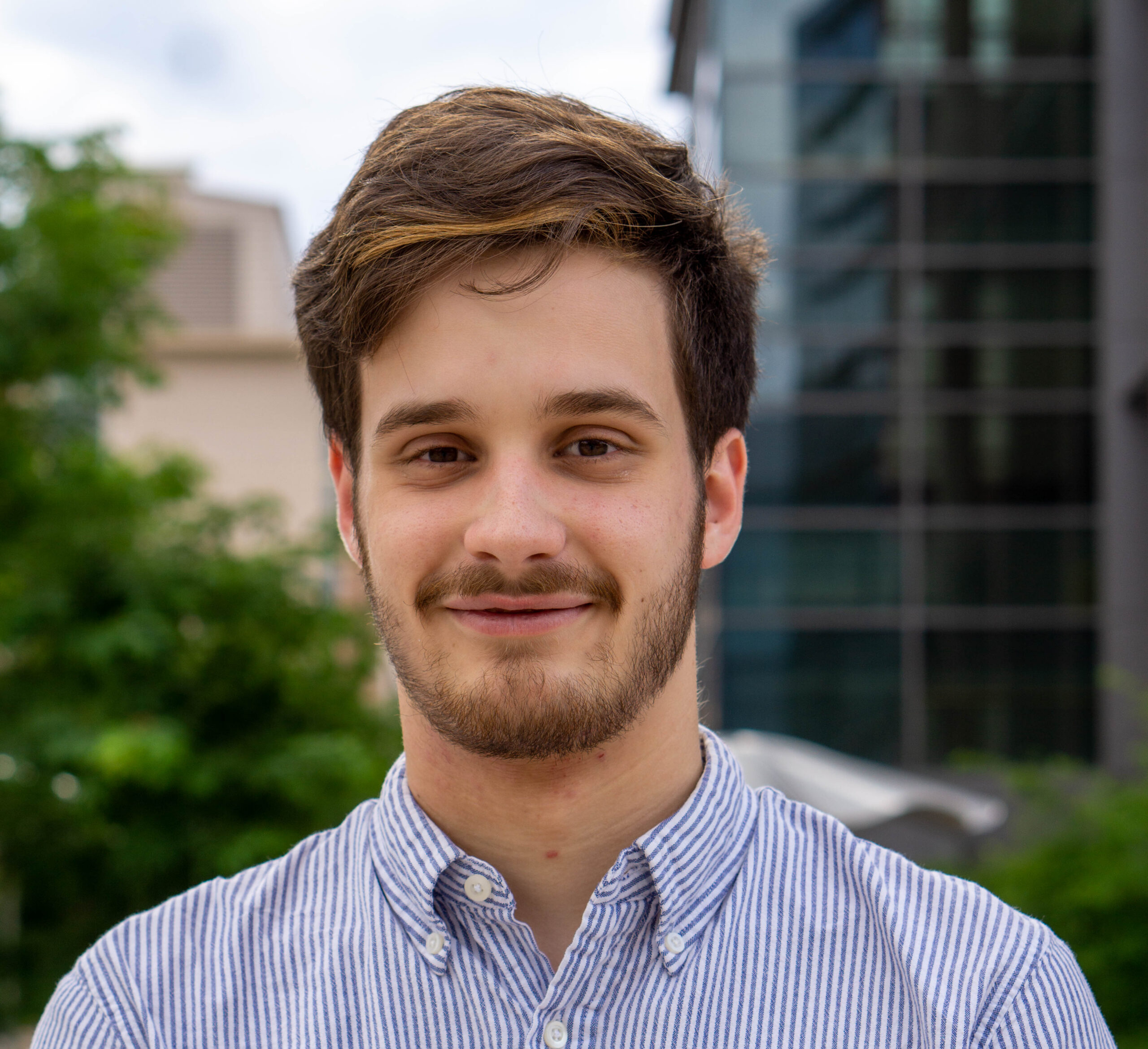 A headshot of a man smiling in front of some blurred buildings